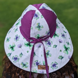 May include: A white sun hat with a purple clover print and a purple ribbon. The hat has a label that says "Kaferme Studio".