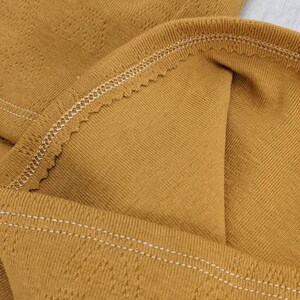 Puede incluir: Primer plano de una tela de color amarillo mostaza con un sutil patr&oacute;n texturizado. La tela tiene una costura blanca cosida a lo largo del borde.