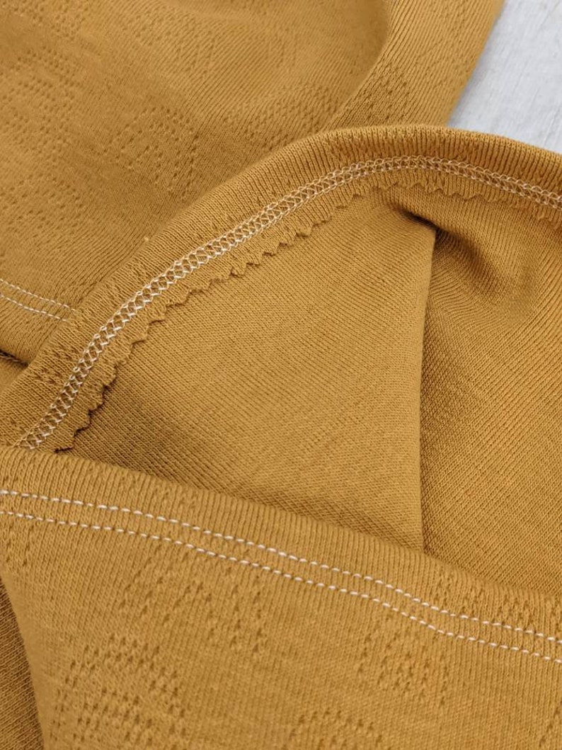 Puede incluir: Primer plano de una tela de color amarillo mostaza con un sutil patr&oacute;n texturizado. La tela tiene una costura blanca cosida a lo largo del borde.