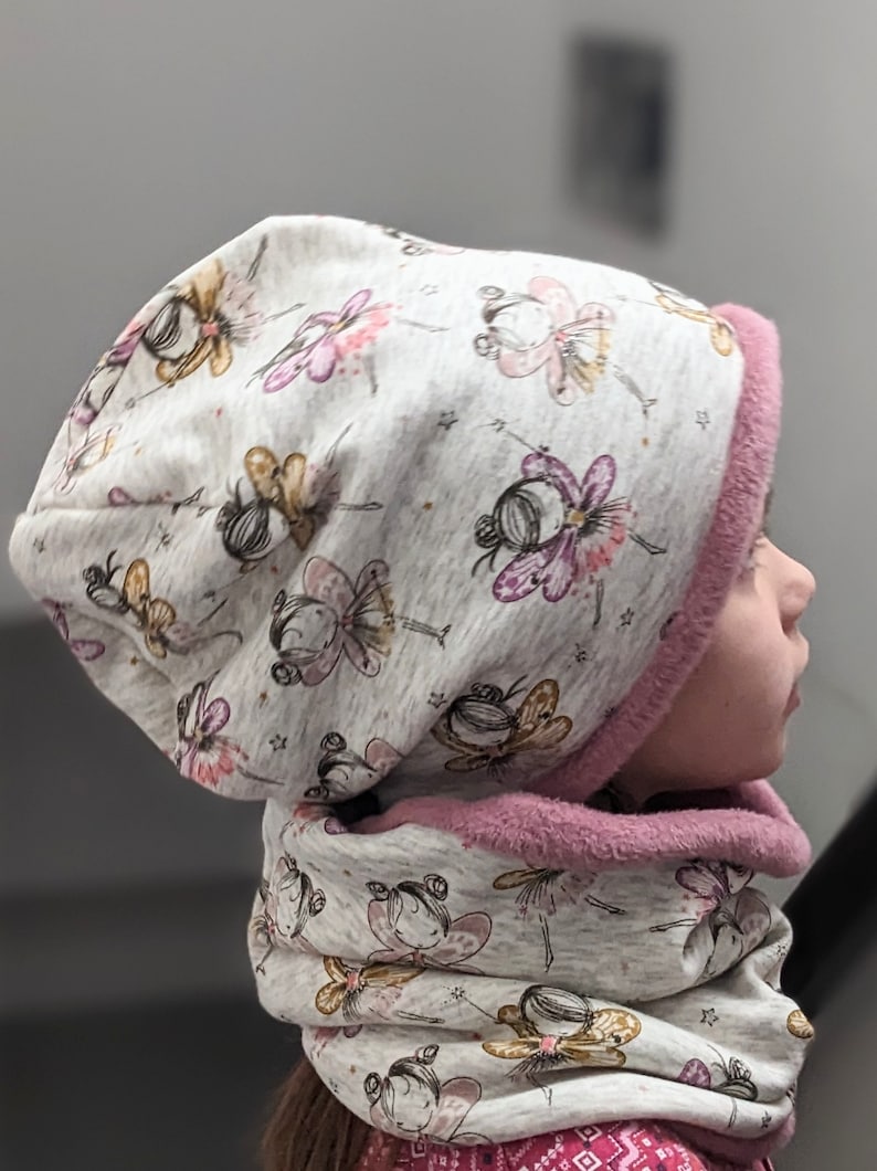 Puede incluir: Un gorro y una bufanda a juego de punto gris y rosa con un patr&oacute;n de hadas con alas y estrellas. El gorro tiene un forro de borreguito rosa.