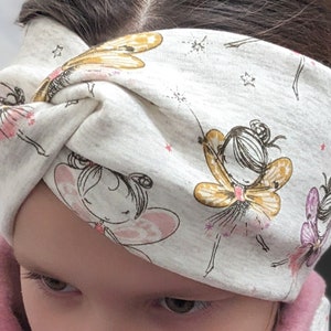 Puede incluir: Una diadema blanca con fondo gris y un patr&oacute;n de hadas con alas y detalles dorados. La diadema est&aacute; anudada en la parte superior de la cabeza.