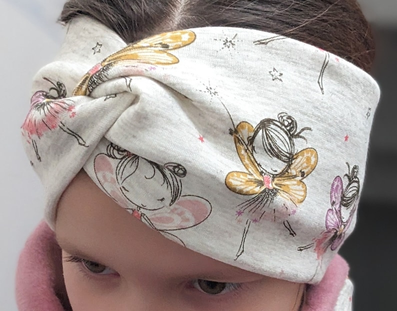 Puede incluir: Una diadema blanca con fondo gris y un patr&oacute;n de hadas con alas y detalles dorados. La diadema est&aacute; anudada en la parte superior de la cabeza.