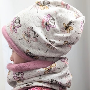 Puede incluir: Un gorro de punto gris y blanco y un cuello a juego con un patr&oacute;n de hadas y estrellas. El gorro tiene un forro de forro polar rosa.