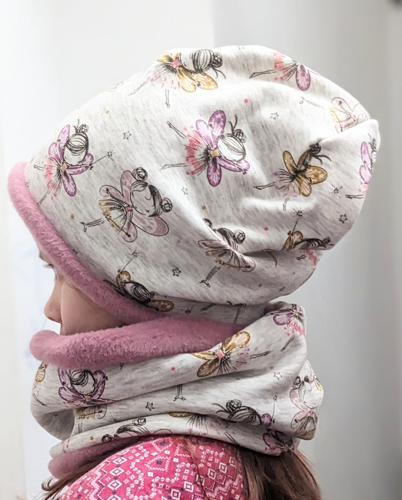Puede incluir: Un gorro de punto gris y blanco y un cuello a juego con un patr&oacute;n de hadas y estrellas. El gorro tiene un forro de forro polar rosa.