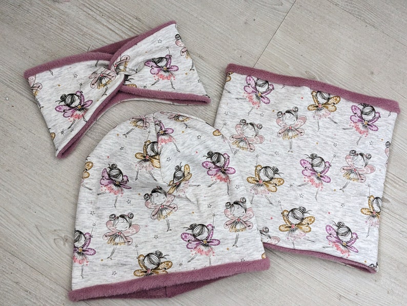 Puede incluir: Un conjunto de tres accesorios de invierno que incluye un gorro gris, un cuello de lana gris y una diadema gris. Los tres art&iacute;culos tienen un patr&oacute;n de hadas con alas rosas y amarillas sobre un fondo gris.