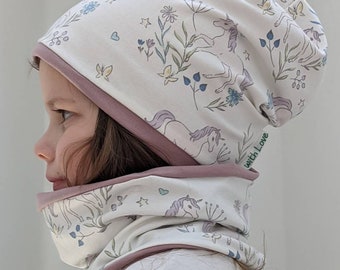 Puede incluir: Conjunto de gorro y cuello de punto blanco con un patrón de unicornio y flores. El gorro tiene un ribete rosa y el cuello tiene un forro rosa. El gorro tiene una etiqueta que dice "with Love".