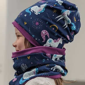 May include: A navy blue beanie and neck warmer set with a whimsical unicorn pattern. The fabric features white unicorns, rainbows, stars, and musical notes. The set has a deep purple trim, creating a contrasting border.