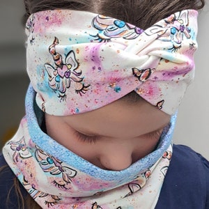 Op de afbeelding: Witte hoofdband en nekwarmer set met een eenhoorn ontwerp. De stof heeft een pastel aquareleffect met roze, blauwe en paarse tinten en gouden glitteraccenten. De eenhoorn heeft een hartvormige neus en een bloemenmanen.