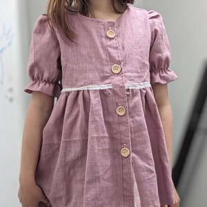 Puede incluir: Una blusa abotonada de color rosa polvoriento con mangas cortas con volantes y un ribete de encaje blanco. La blusa presenta grandes botones de madera y está hecha de una tela texturizada. La prenda forma parte de una colección de ropa infantil.
