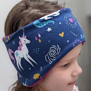May include: A navy blue headband featuring a white unicorn with a pink mane and tail, surrounded by colorful stars, hearts, and musical notes. The headband is lined with a pink fabric and is designed for comfort and style.