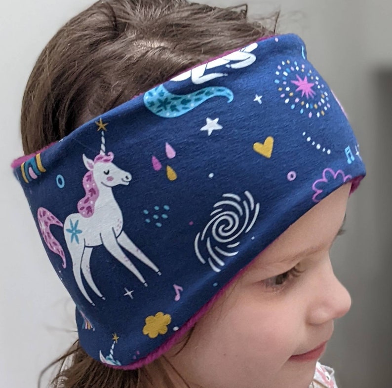 May include: A navy blue headband featuring a white unicorn with a pink mane and tail, surrounded by colorful stars, hearts, and musical notes. The headband is lined with a pink fabric and is designed for comfort and style.