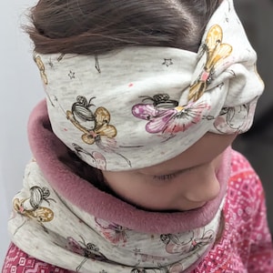 Puede incluir: Una diadema gris y blanca con un dise&ntilde;o anudado y un patr&oacute;n de hadas y mariposas. La diadema la lleva puesta un ni&ntilde;o con el pelo casta&ntilde;o. El ni&ntilde;o tambi&eacute;n lleva un cuello de lana gris y blanco con forro de borreguito rosa.