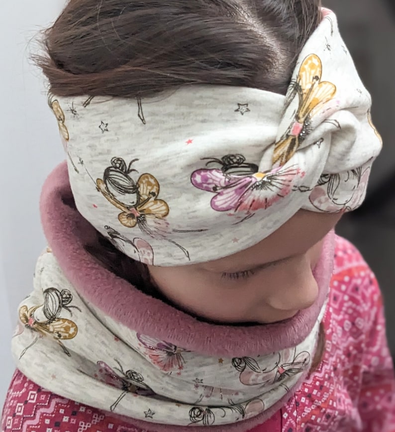 Puede incluir: Una diadema gris y blanca con un dise&ntilde;o anudado y un patr&oacute;n de hadas y mariposas. La diadema la lleva puesta un ni&ntilde;o con el pelo casta&ntilde;o. El ni&ntilde;o tambi&eacute;n lleva un cuello de lana gris y blanco con forro de borreguito rosa.