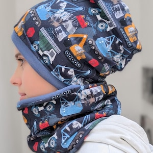 May include: A blue and black knit beanie and neck warmer set with a construction vehicle print. The beanie has a fold-over cuff and the neck warmer is a loop style.