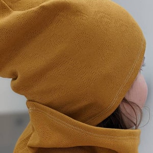 Puede incluir: Un conjunto de gorro y cuello de punto amarillo mostaza. El gorro tiene un patr&oacute;n texturizado y el cuello est&aacute; doblado hacia abajo.
