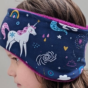 May include: A navy blue ear warmer featuring a unicorn, rainbows, stars, and other celestial designs. The interior is lined with a vibrant pink plush material. This headband is designed for warmth and comfort.