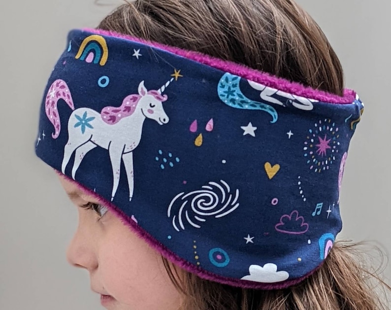 May include: A navy blue ear warmer featuring a unicorn, rainbows, stars, and other celestial designs. The interior is lined with a vibrant pink plush material. This headband is designed for warmth and comfort.