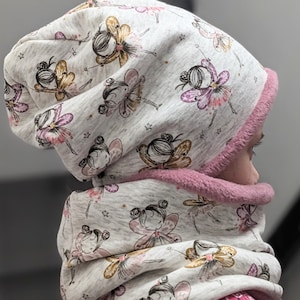 Könnte beinhalten: Eine graue und weiße Strickmütze und ein passender Schal mit einem Muster aus Feen mit rosa Flügeln. Die Mütze hat ein rosa Fleecefutter.