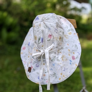 Op de afbeelding: Een witte zonnehoed met een bloemenprint en een witte strik. De hoed is gemaakt van een lichtgewicht stof en heeft een brede rand.