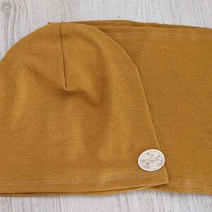 Puede incluir: Un conjunto de gorro y cuello de punto a juego de color mostaza. El gorro tiene una peque&ntilde;a etiqueta blanca con un dise&ntilde;o.
