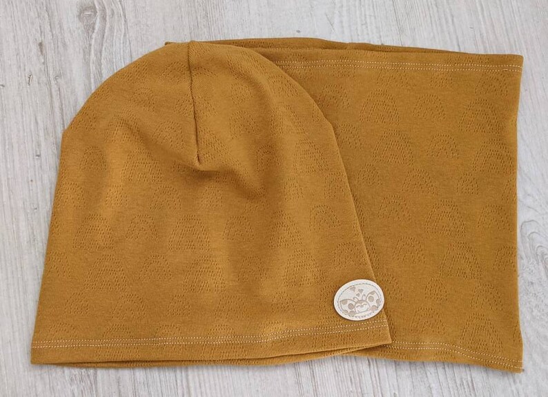 Puede incluir: Un conjunto de gorro y cuello de punto a juego de color mostaza. El gorro tiene una peque&ntilde;a etiqueta blanca con un dise&ntilde;o.