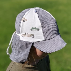 May include: A gray corduroy sun hat with a white panel featuring a vintage car print. The hat has a wide brim and ear flaps.