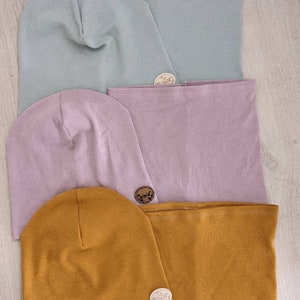 Puede incluir: Tres juegos de gorros y cuellos de punto a juego en azul claro, lavanda y amarillo mostaza. Cada juego tiene una peque&ntilde;a etiqueta de madera con un dise&ntilde;o.