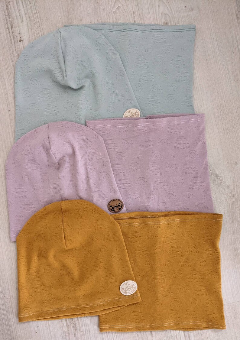 Puede incluir: Tres juegos de gorros y cuellos de punto a juego en azul claro, lavanda y amarillo mostaza. Cada juego tiene una peque&ntilde;a etiqueta de madera con un dise&ntilde;o.