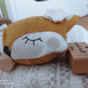 May include: A stuffed fox toy with a mustard yellow head and white face, embroidered with closed eyes. The fox has a white frill and a black nose. A wooden stamp with the text "made with love" is next to the toy.