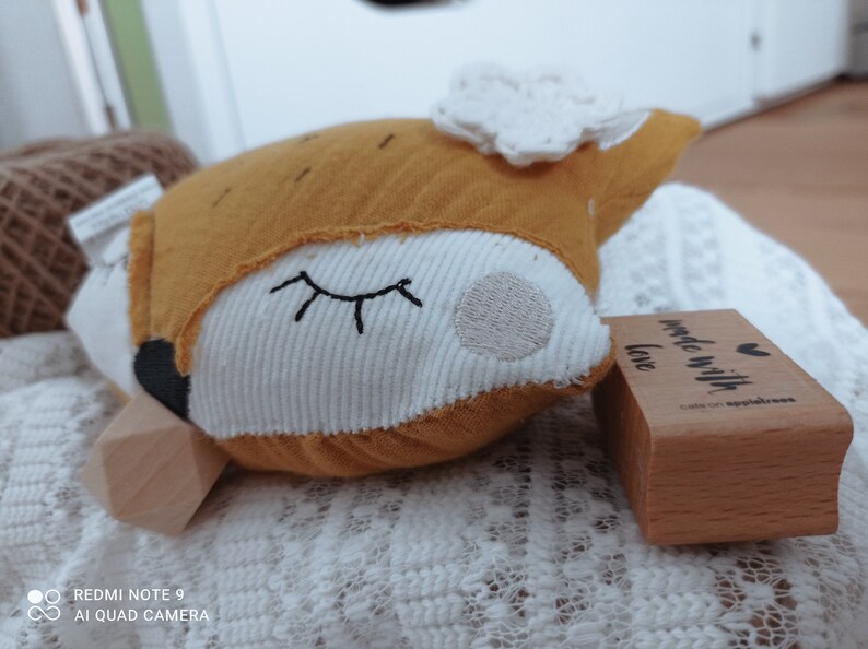 May include: A stuffed fox toy with a mustard yellow head and white face, embroidered with closed eyes. The fox has a white frill and a black nose. A wooden stamp with the text "made with love" is next to the toy.