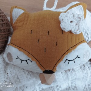 May include: A plush fox-shaped baby toy with a mustard-yellow face, white accents, and a wooden teether. The fox has embroidered eyes and a white crocheted flower. A tag reads "made with love".