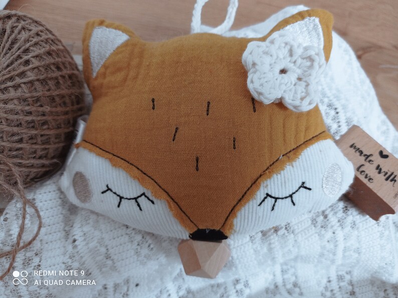May include: A plush fox-shaped baby toy with a mustard-yellow face, white accents, and a wooden teether. The fox has embroidered eyes and a white crocheted flower. A tag reads "made with love".