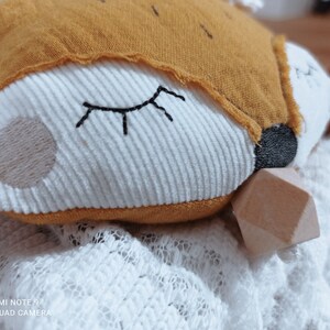 May include: A plush fox-shaped baby toy with a white corduroy face, embroidered closed eyes, and a tan embroidered cheek. The fox has a mustard-colored hood and a wooden geometric shape attached. The toy rests on a white lace fabric.
