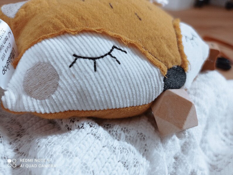 May include: A plush fox-shaped baby toy with a white corduroy face, embroidered closed eyes, and a tan embroidered cheek. The fox has a mustard-colored hood and a wooden geometric shape attached. The toy rests on a white lace fabric.