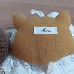 May include: Mustard-colored fox-shaped plush toy with a white label that reads "Villala". The toy has a loop for hanging and is placed on a white lace fabric. A ball of twine is visible in the background.