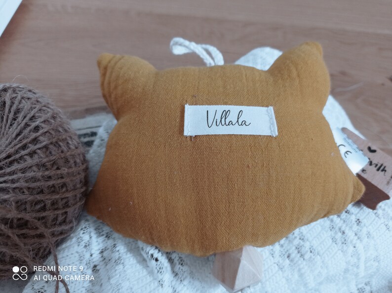 May include: Mustard-colored fox-shaped plush toy with a white label that reads "Villala". The toy has a loop for hanging and is placed on a white lace fabric. A ball of twine is visible in the background.