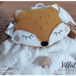 May include: A plush fox-shaped baby mobile in mustard yellow and white, with closed eyes and a wooden geometric pendant. The mobile rests on a white lace blanket, with a ball of twine and a stamp that says "made with love". The text "Villala" is visible.