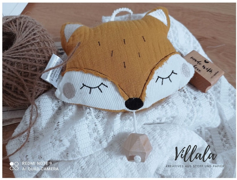 May include: A plush fox-shaped baby mobile in mustard yellow and white, with closed eyes and a wooden geometric pendant. The mobile rests on a white lace blanket, with a ball of twine and a stamp that says "made with love". The text "Villala" is visible.