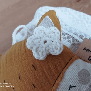 May include: A handmade fox-shaped plush toy with a white crocheted flower accent. The fox is primarily orange with white and silver details. The toy rests on a white lace fabric, with a wooden stamp that reads "made with love".