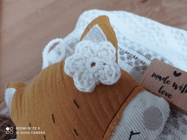 May include: A handmade fox-shaped plush toy with a white crocheted flower accent. The fox is primarily orange with white and silver details. The toy rests on a white lace fabric, with a wooden stamp that reads "made with love".