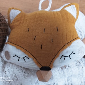 May include: A plush fox-shaped baby toy with a mustard-yellow face, white accents, and closed eyes. The toy has a wooden teether at the bottom and a loop for hanging. The words "made with love" are on a small wooden tag.