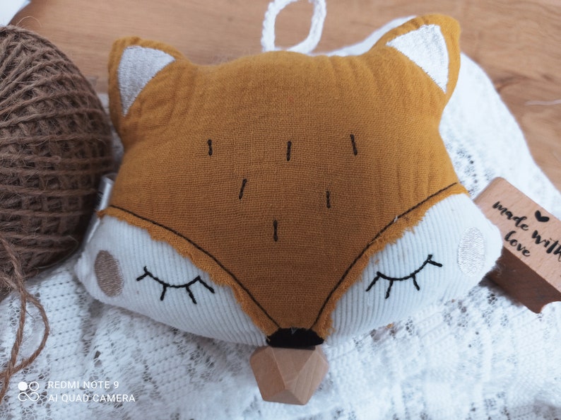 May include: A plush fox-shaped baby toy with a mustard-yellow face, white accents, and closed eyes. The toy has a wooden teether at the bottom and a loop for hanging. The words "made with love" are on a small wooden tag.