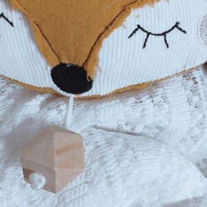 May include: A plush fox-shaped toy with a mustard-colored face and white accents. The fox has embroidered eyes and a black nose. A small wooden geometric shape hangs below on a white string. The toy rests on a white lace surface.