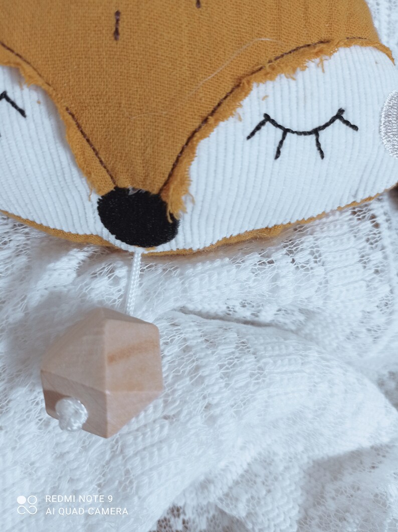 May include: A plush fox-shaped toy with a mustard-colored face and white accents. The fox has embroidered eyes and a black nose. A small wooden geometric shape hangs below on a white string. The toy rests on a white lace surface.