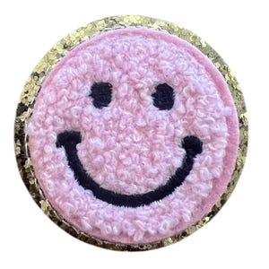 Könnte beinhalten: Ein rosa Smiley-Aufnäher mit einer strukturierten, flauschigen Oberfläche und schwarzen Augen und Mund. Der runde Aufnäher ist von einem goldenen Glitzerrahmen umgeben. Dieser Aufnäher kann zum Dekorieren von Kleidung oder Accessoires verwendet werden.