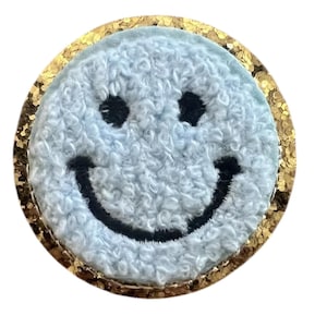 Könnte beinhalten: Ein hellblaues Smiley-Gesicht-Patch mit einem strukturierten, flauschigen Aussehen. Das Smiley-Gesicht hat schwarze Augen und einen schwarzen, gebogenen Mund. Das Patch ist von einem goldenen Glitzerrahmen umgeben. Das Patch ist rund.