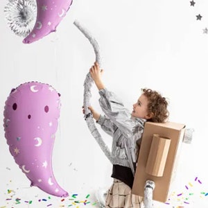 Puede incluir: Un ni&ntilde;o con un disfraz de cohete de cart&oacute;n sostiene una serpentina plateada unida a dos globos morados con un patr&oacute;n de luna y estrellas. El ni&ntilde;o est&aacute; de pie sobre una superficie blanca cubierta de confeti de colores.