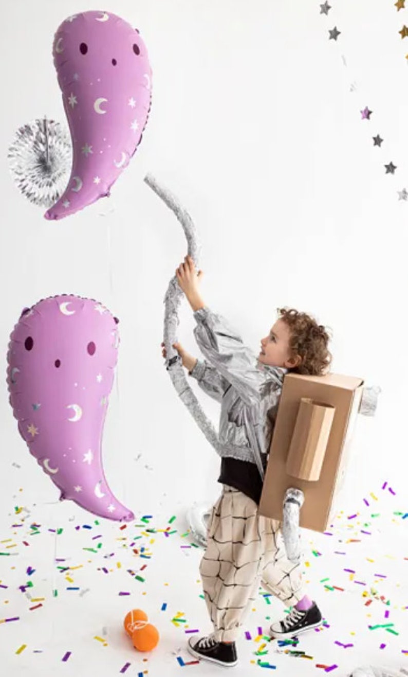 Puede incluir: Un ni&ntilde;o con un disfraz de cohete de cart&oacute;n sostiene una serpentina plateada unida a dos globos morados con un patr&oacute;n de luna y estrellas. El ni&ntilde;o est&aacute; de pie sobre una superficie blanca cubierta de confeti de colores.