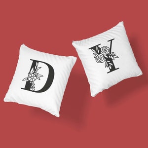Może przedstawiać: Dwie białe poszewki na poduszki z czarnymi wzorami liter kwiatowych. Poduszki przedstawiają litery "D" i "Y" z kwiatowymi akcentami.