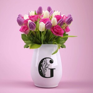 Może przedstawiać: Biały ceramiczny wazon z czarnym wzorem kwiatowym i literą "G" z przodu. Wazon jest wypełniony bukietem r&oacute;żowych, białych i fioletowych tulipan&oacute;w.